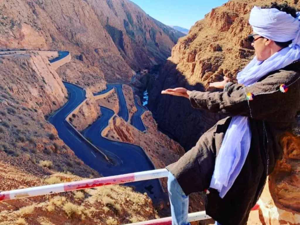 Guide in traditional robes with arms outstretched above the winding road of Dades Gorge, Morocco